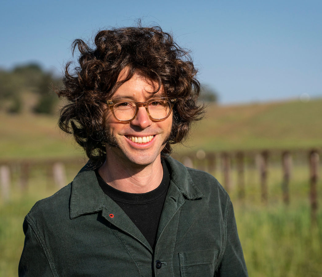 Man with dark curly hair and glasses looks at camera, with vinyards behind him