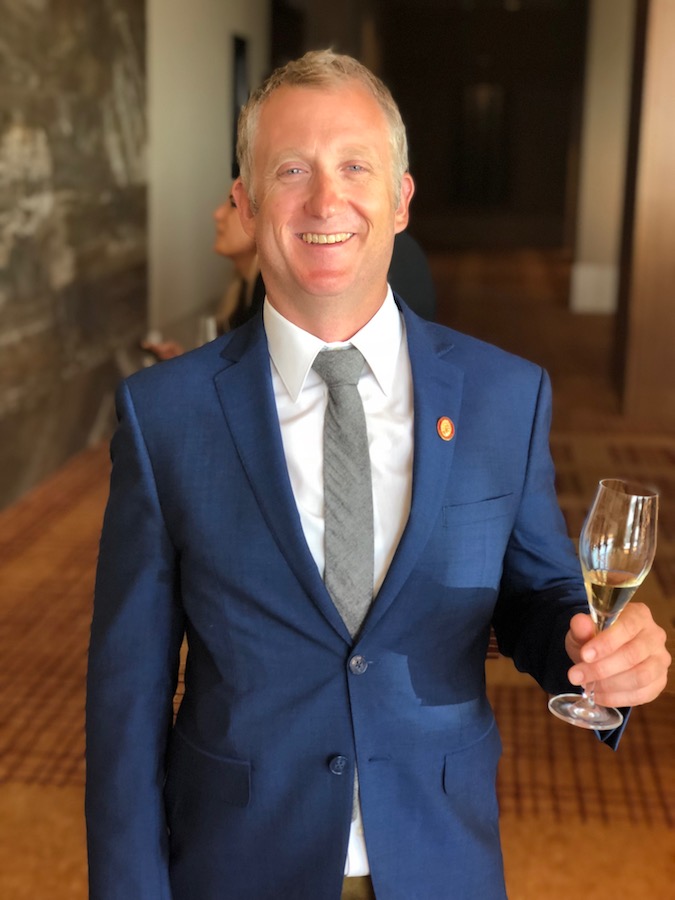 Man in blue suit smiles at the camera and holds Champagne flute