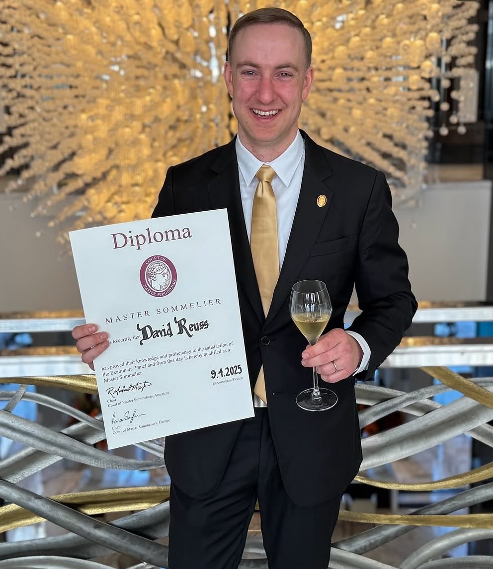 man holding paper diploma and glass of white wine smiles at camera