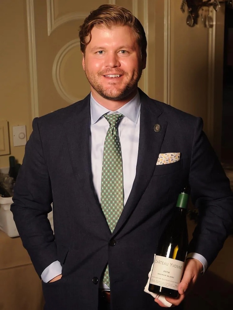 Man with blonde hair in a suit and green tie looks at camera holding bottle of wine and smiling