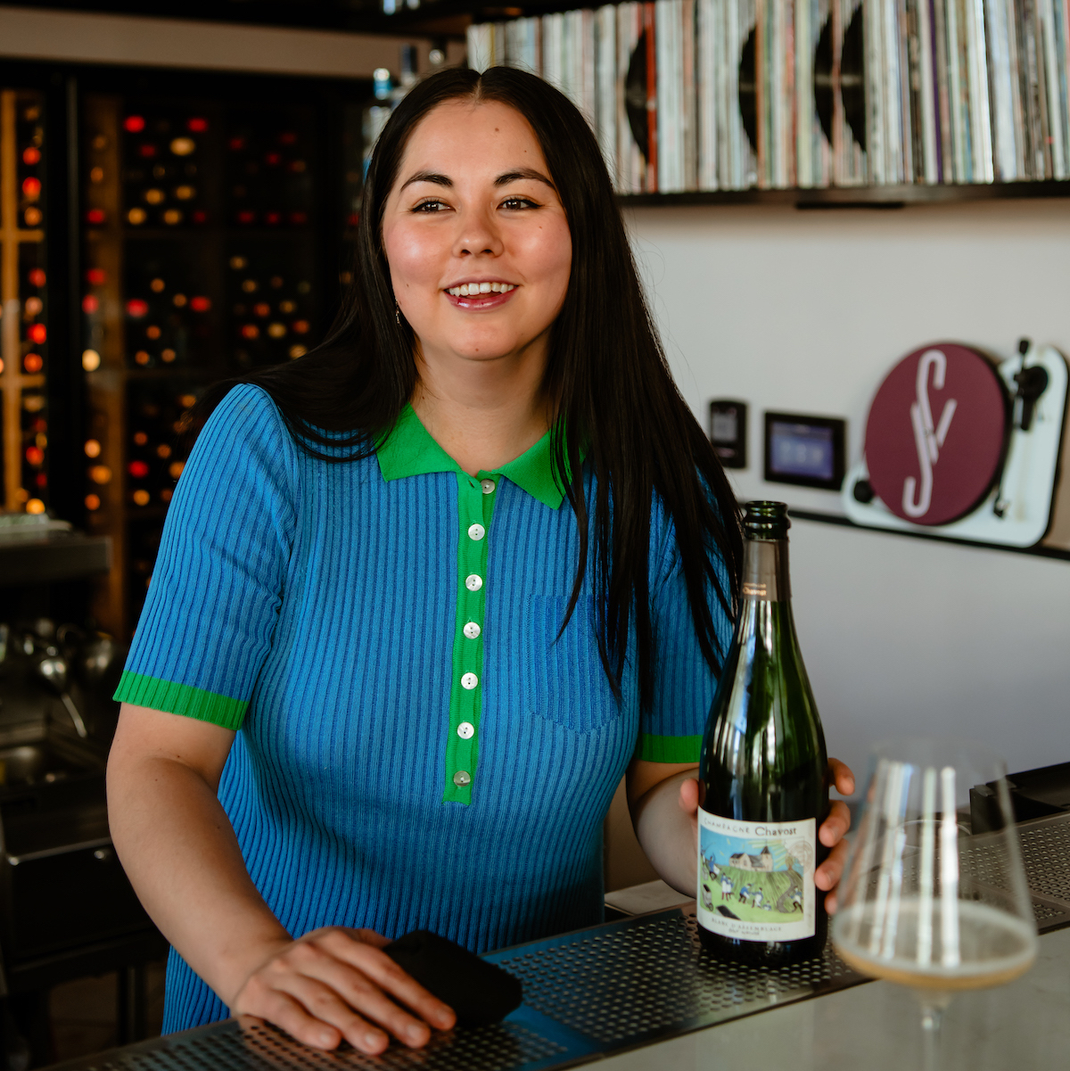 Dark-haired woman wearing teal looks into the distance while smiling with wine bottle in hand and a shelf of records in the background