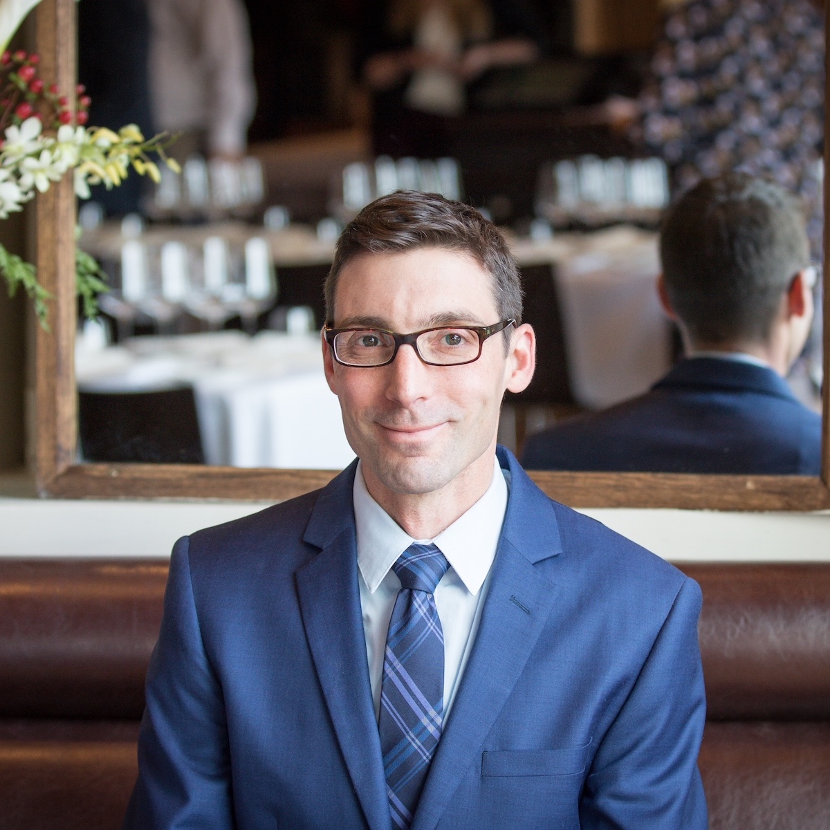 Man in blue suit with dark hair and glasses smiles and looks at the camera in a restaurant booth