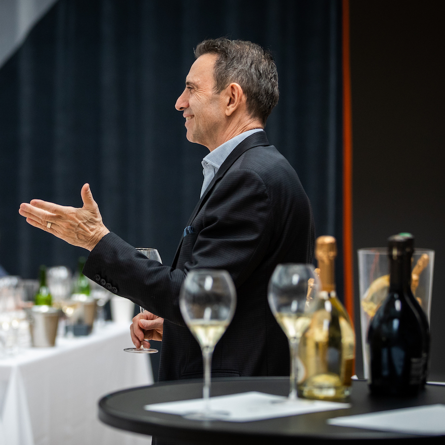 Man in dark suit smiles while speaking with Champagne bottles and glasses in the foreground
