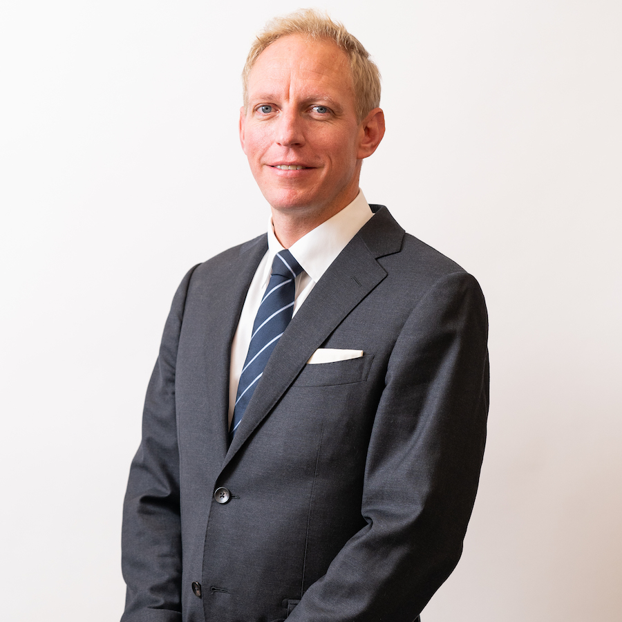 Man in suit with ligh-colored hair stands against a white background and smiles at camera