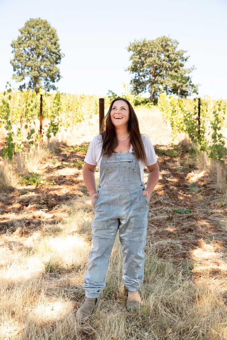 Woman in overalls stands in vineyard and grins looking upward