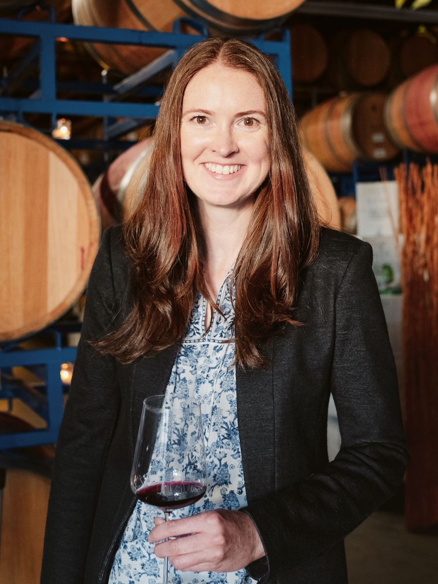 woman standing in front of wine barrels and holding wine glass smiles at the camera