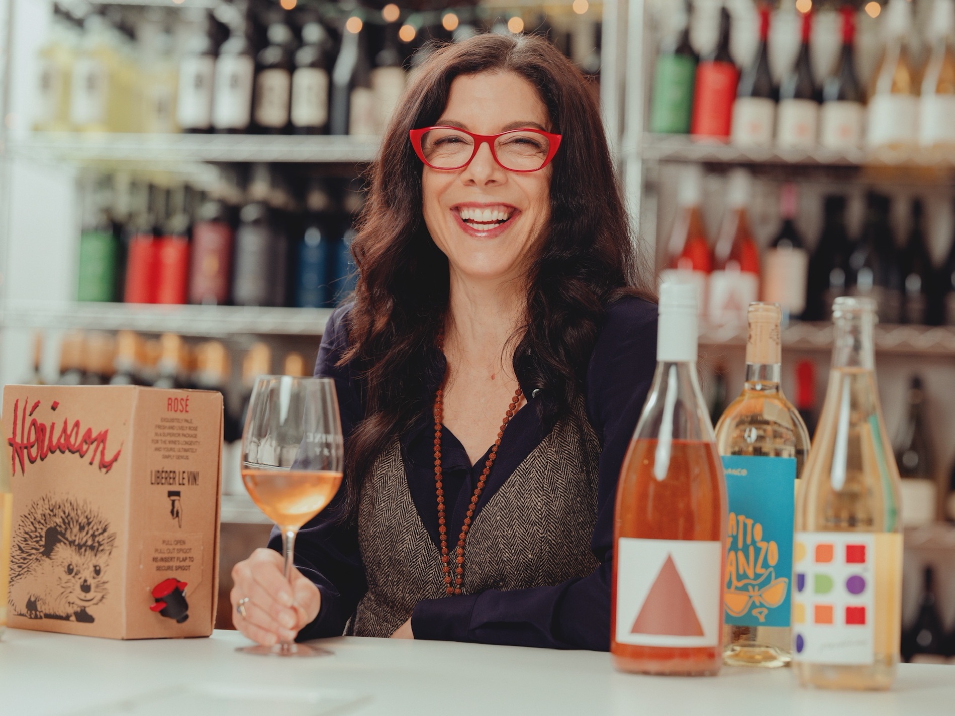 Woman in red glasses looks at camera and smiles, with wine bottles and BiBs in the background