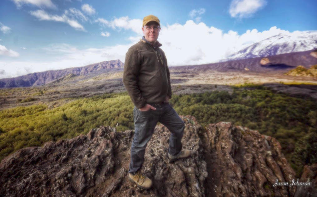 Man standing on volcanic rock looks at camera with mountainous landscape in the backgound