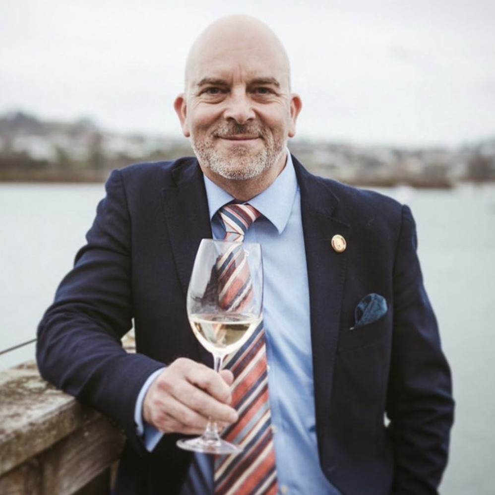 Man in suit holding glass of wine stands at dock and looks toward camera