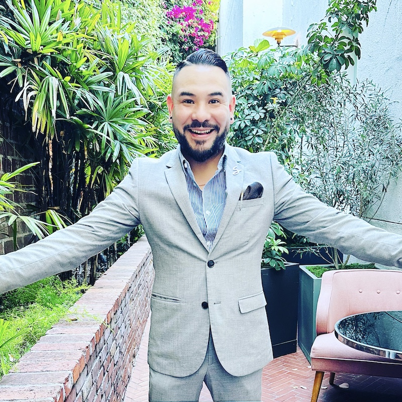 Man in suit smiles at the camera with arms outstretched with beautiful garden in the background