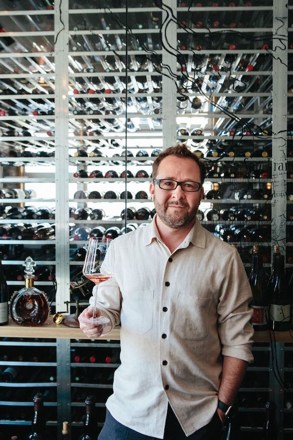 Man stands in front of large glass-enclosed wine cellar holding glass of wine