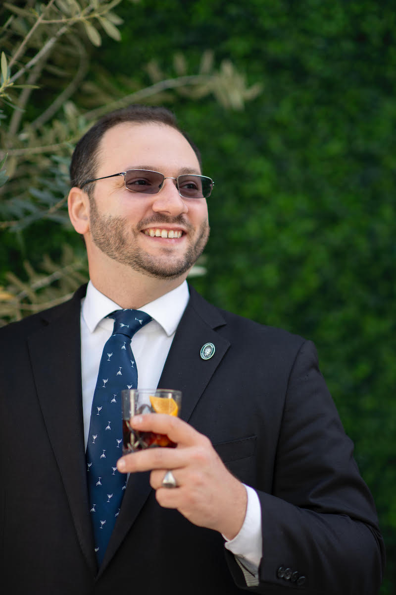 Man in dark suit holds drink and looks into the distance smiling
