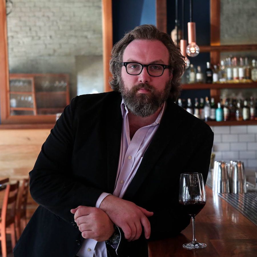 Bearded man in jacket and purple shirt looks intently at the camera while leaning against bar with wine glass