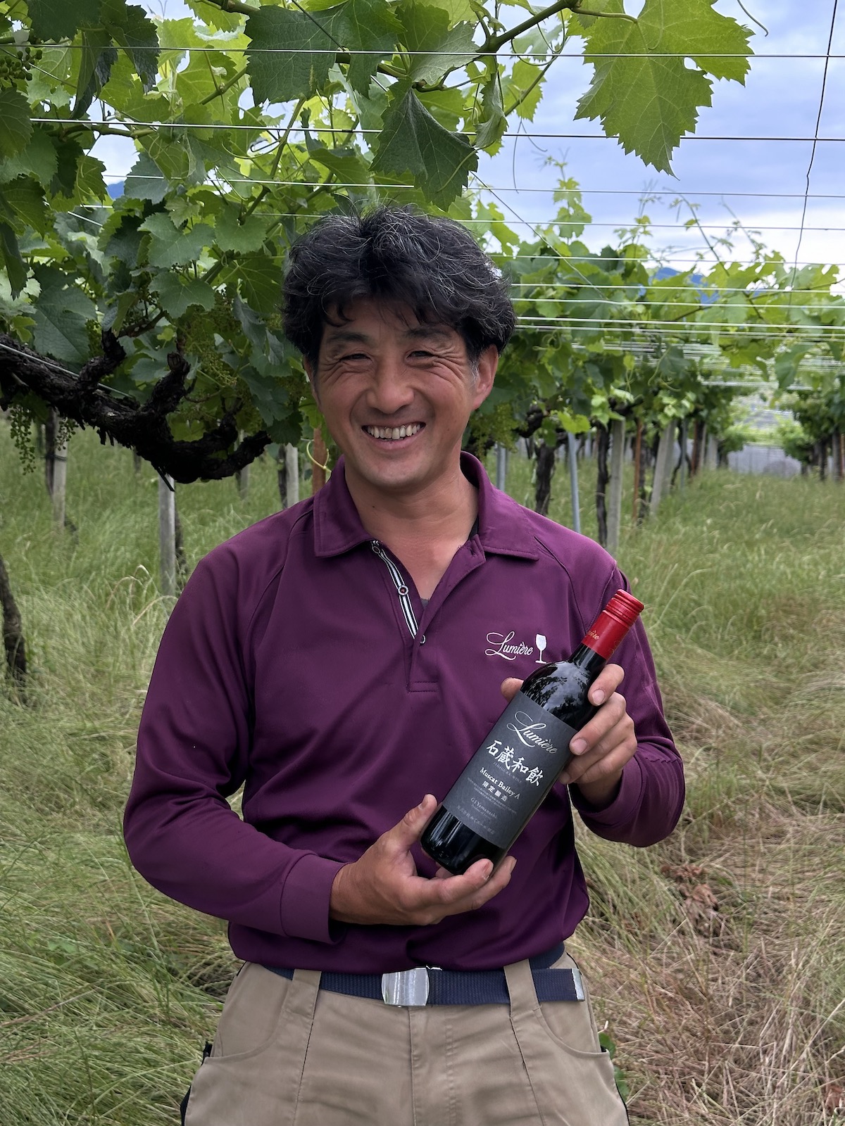 Man in burgundy shirt holds wine bottle in a vineyard