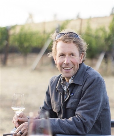 Man in flannel shirt and gray jacket looks past camera while holding a glass of wine and smiling